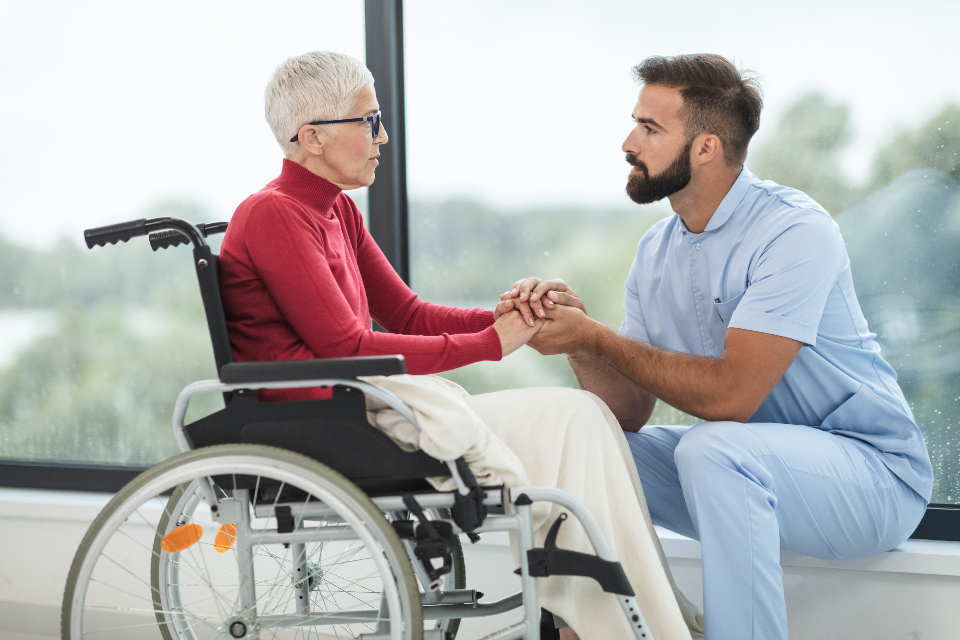 A caregiver sitting beside a client in a wheelchair, holding hands by a bright window
